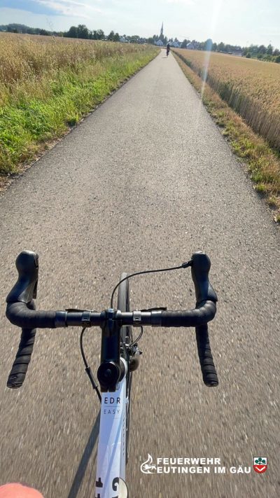 ein Radfahrer bei Sonnenschein auf einem geteerten Feldweg zwischen Weizenfeldern, im Hintergrund ein Dorf